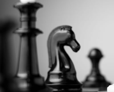 A close up image of a knight chess piece on a chess board. Out of focus in the foreground is the king, and out of focus in the background is a pawn. All three chess pieces are black in colour.